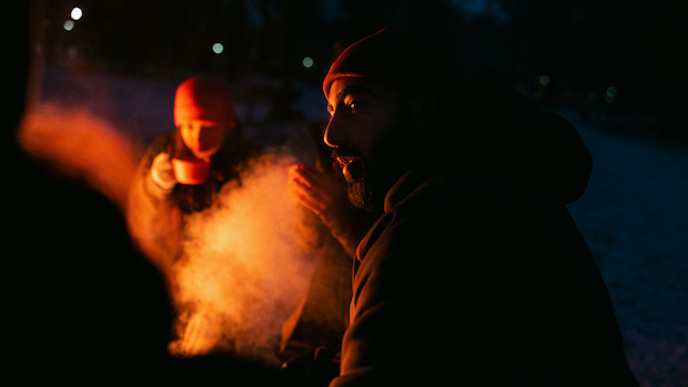 Två personer sitter utomhus med varma kläder på sig. Ena personen dricker något ur en mugg. 