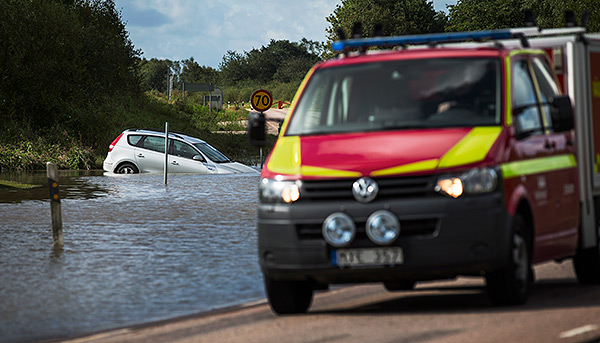 Bilden visar en bil som fastnat på en översvämmad väg. I förgrunden står en brandbil.