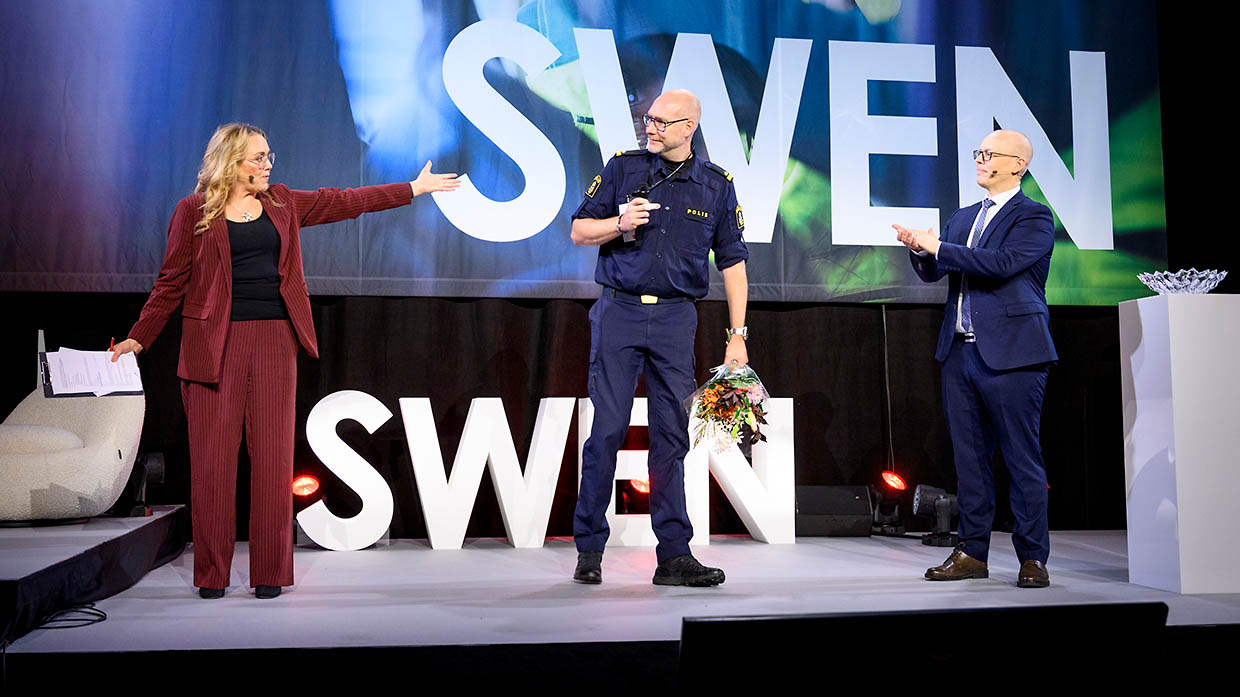 Ellinor Persson, moderator, Fredrik Ryberg, Polisen och Ronny Harpe, MSB står på en scen. Ronny Harpe applåderar. Fredrik Ryberg har en blombukett i handen.