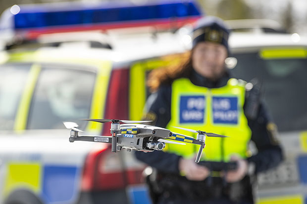Polis i uniform och varselväst styr en flygande drönare med hjälp av en fjärrkontroll som hon håller i händerna.